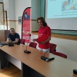 Μια γυναίκα με κόκκινη μπλούζα του συλλόγου στέκεται πίσω από το βήμα, κρατώντας χαρτιά, ενώ ο ομιλητής κάθεται δίπλα της.