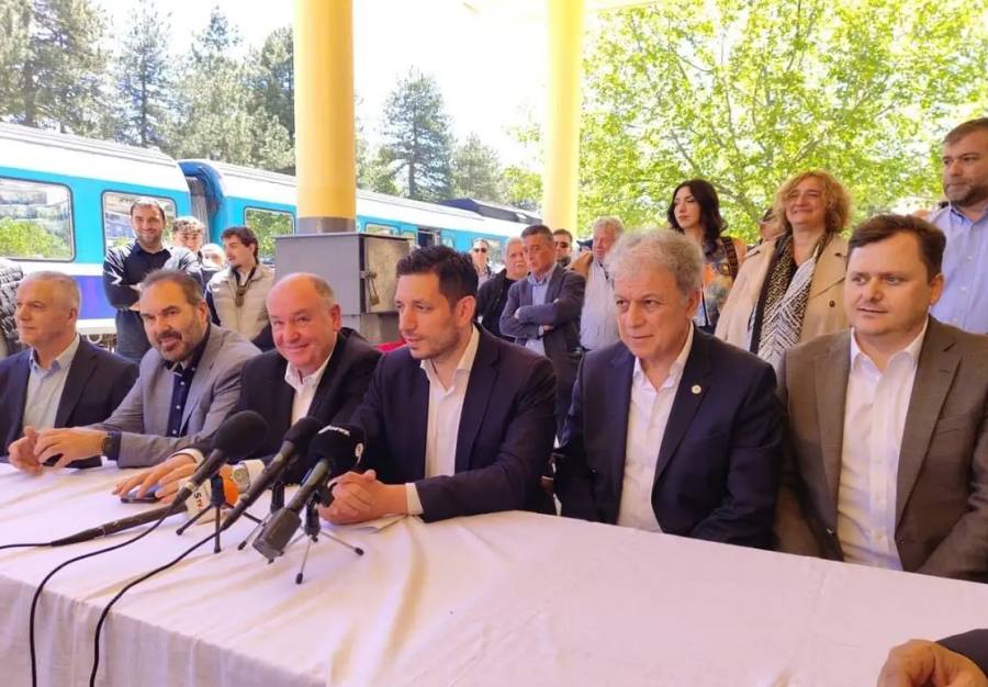 Ομάδα πολιτικών ανδρών σε συνέντευξη Τύπου μπροστά σε τραπέζι με μικρόφωνα, με τρένο ορατό στο βάθος.